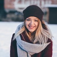 A smiling young woman with blonde hair wearing a dark knit beanie, a thick light gray scarf, and a maroon coat, standing outdoors in a snowy setting.