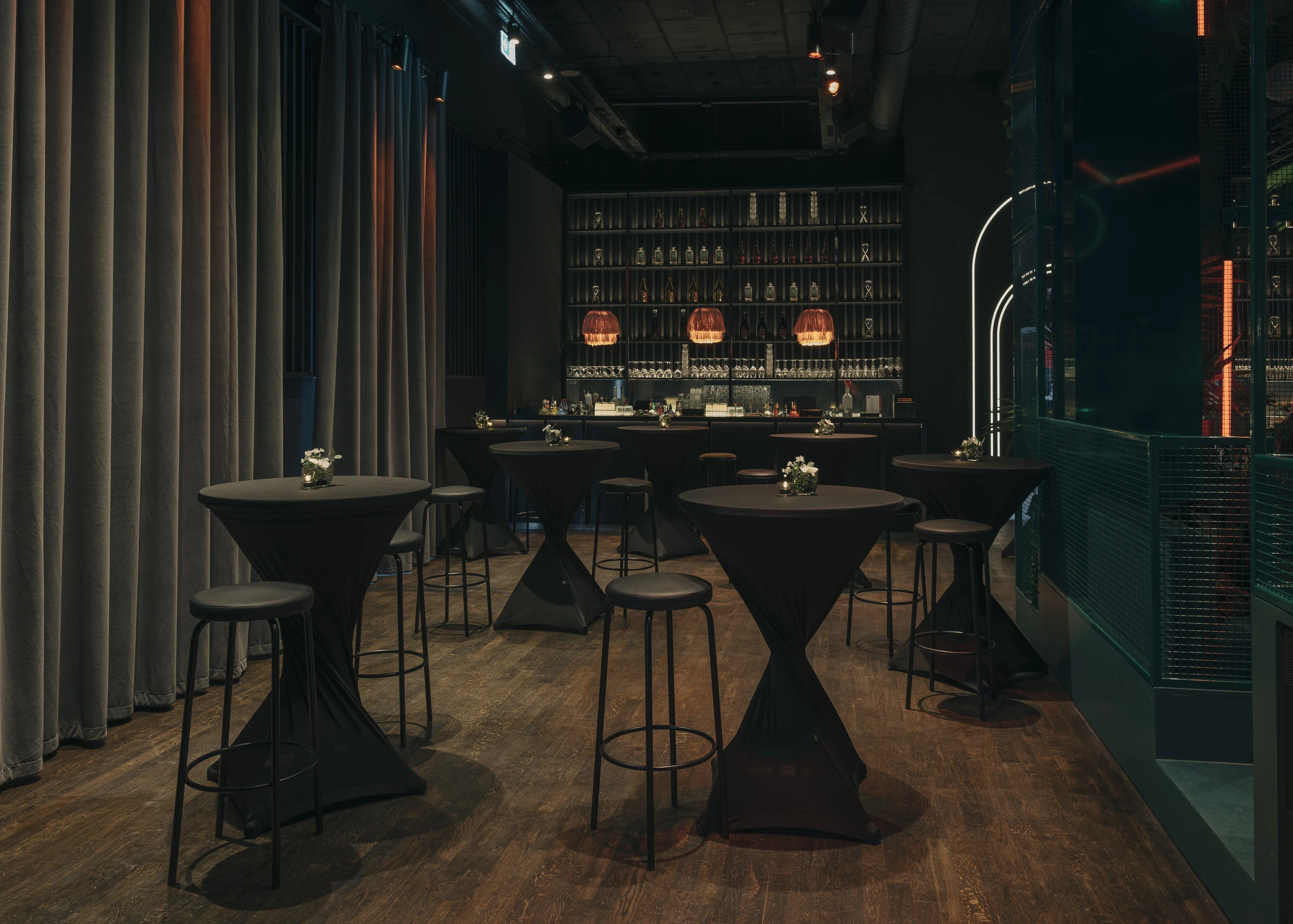 Bar area at NATE CLUB with black draped high tables and stools, grey velvet curtains and an illuminated back bar with copper wire pendant lights and bottle shelves.