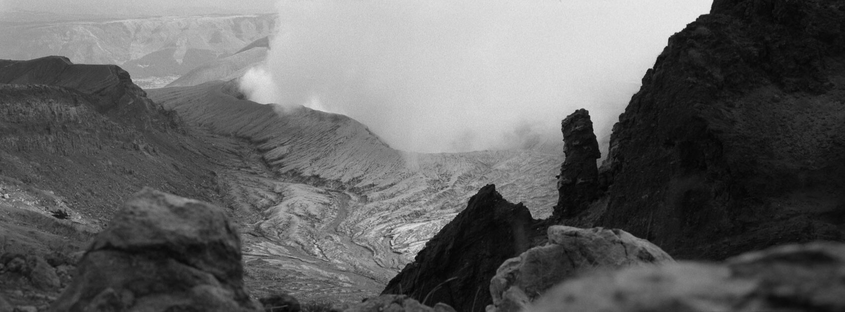 Japan hiking at Mount Aso: volcanic crater with steam clouds, with a area at the front that is cast in shadows whilst the steam is bright creating a dramatic black-and-white landscape, captured on Ilford film in Kyushu's otherworldly terrain.