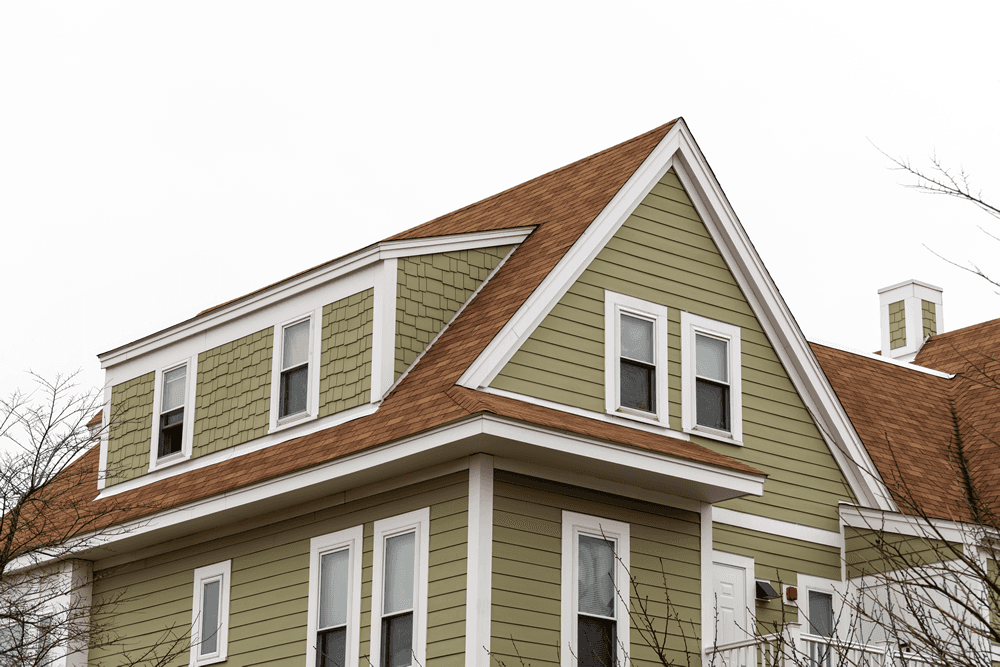 A close-up view of a house with sage-green siding and white trim, featuring a steeply pitched roof covered in brown shingles. The architectural details include a mix of clapboard and shake siding, multiple windows with white frames, and sharp gables that create a classic, symmetrical look.