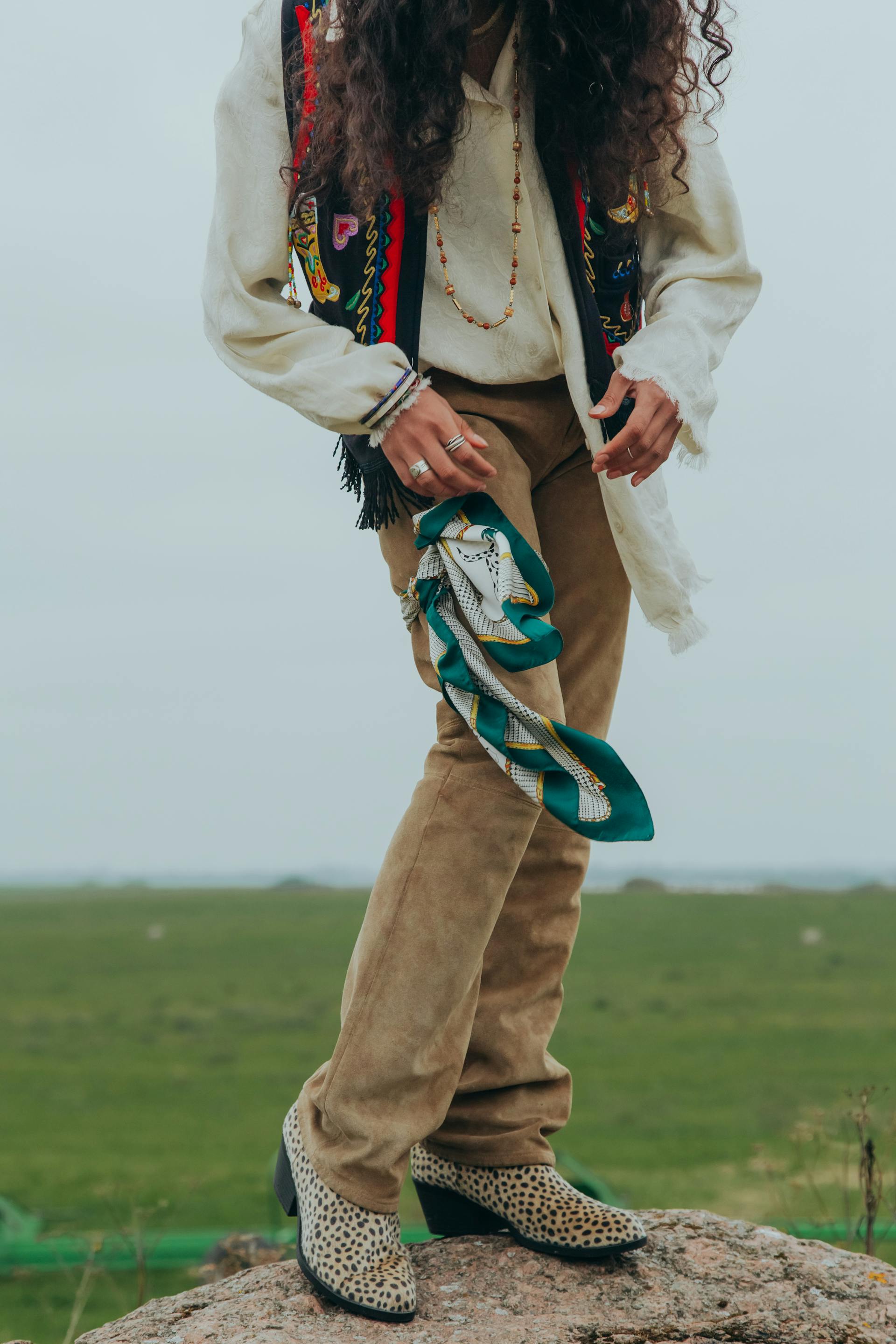 A person wearing leopard-print boots and an embroidered vest stands on a rock in a field