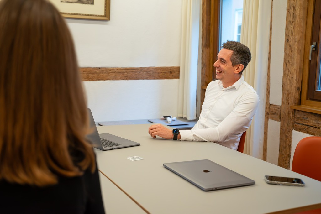 Eine Person sitzt an einem Tisch in einem Besprechungsraum und blickt auf eine andere Person, die mit dem Rücken zur Kamera s