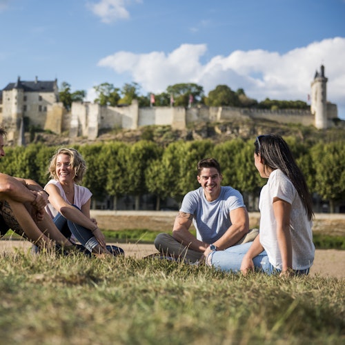 Cuatro personas se sientan y charlan sobre el césped con un edificio histórico y árboles al fondo, bajo un cielo azul con algunas nubes.