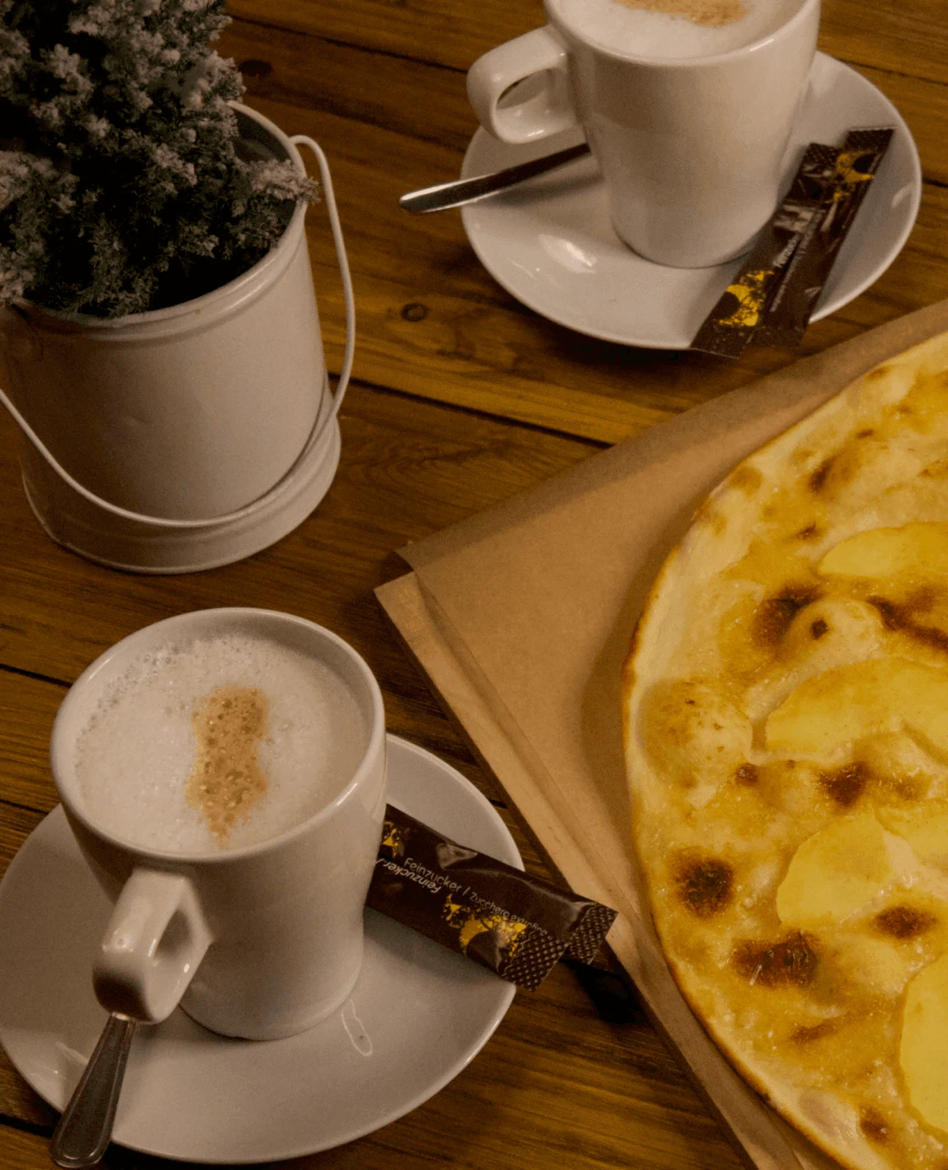 Flammkuchen mit Apfelbelag auf einem Holzbrett, daneben zwei Tassen Cappuccino und eine kleine Deko-Pflanze.