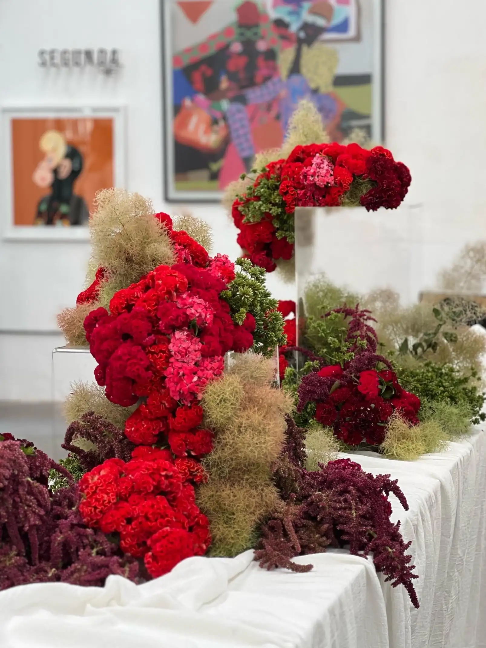Instalación floral contemporánea con flores rojas y fucsias sobre mesas blancas para galerías y artistas que buscan un montaje botánico expresivo y de alto impacto visual.