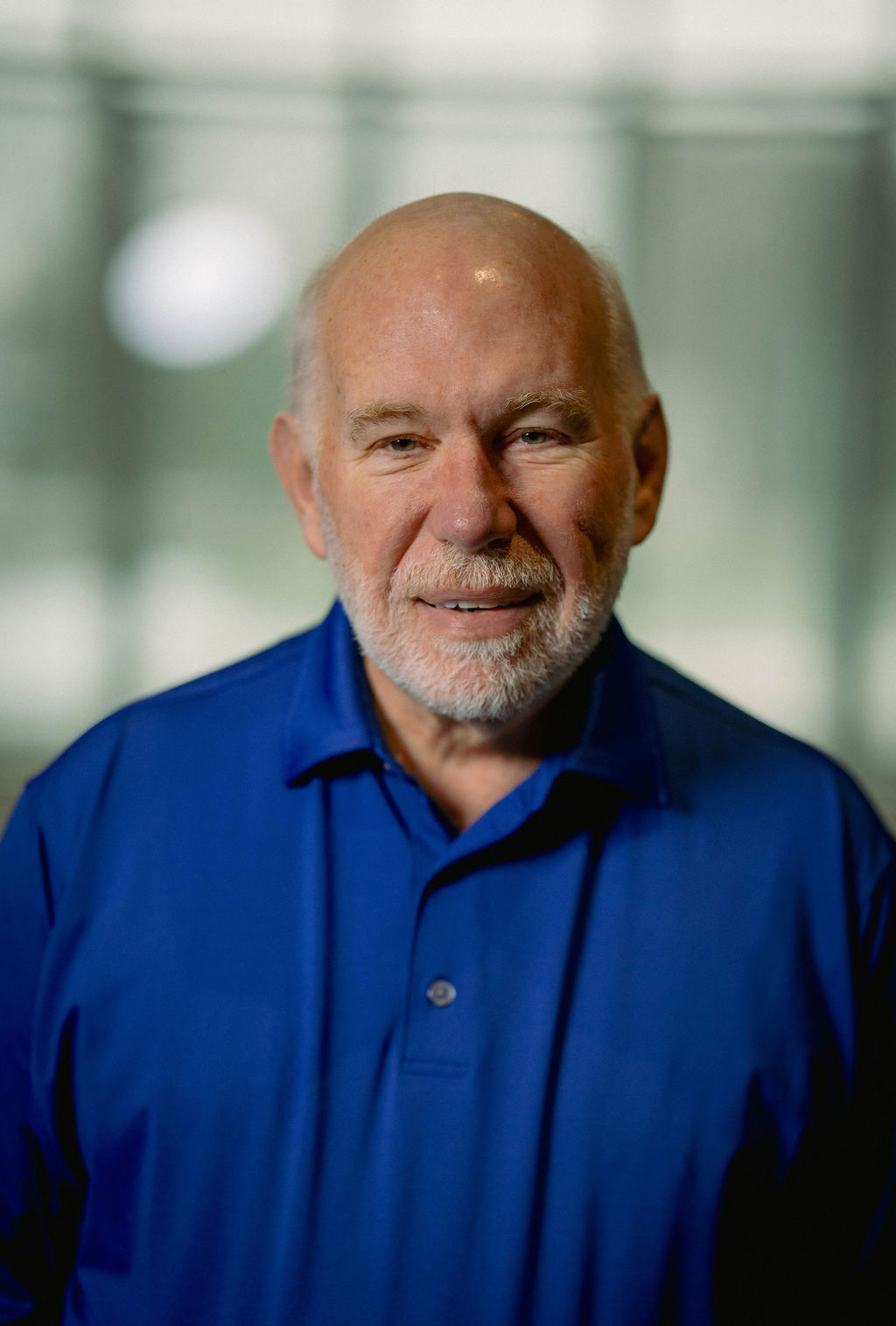Portrait of a young man in a brown coat and beige turtleneck, with light brown hair and a calm expression against a gray background.
