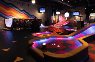 Smiling young woman with long hair standing against a dark green background, holding a finger to her chin.