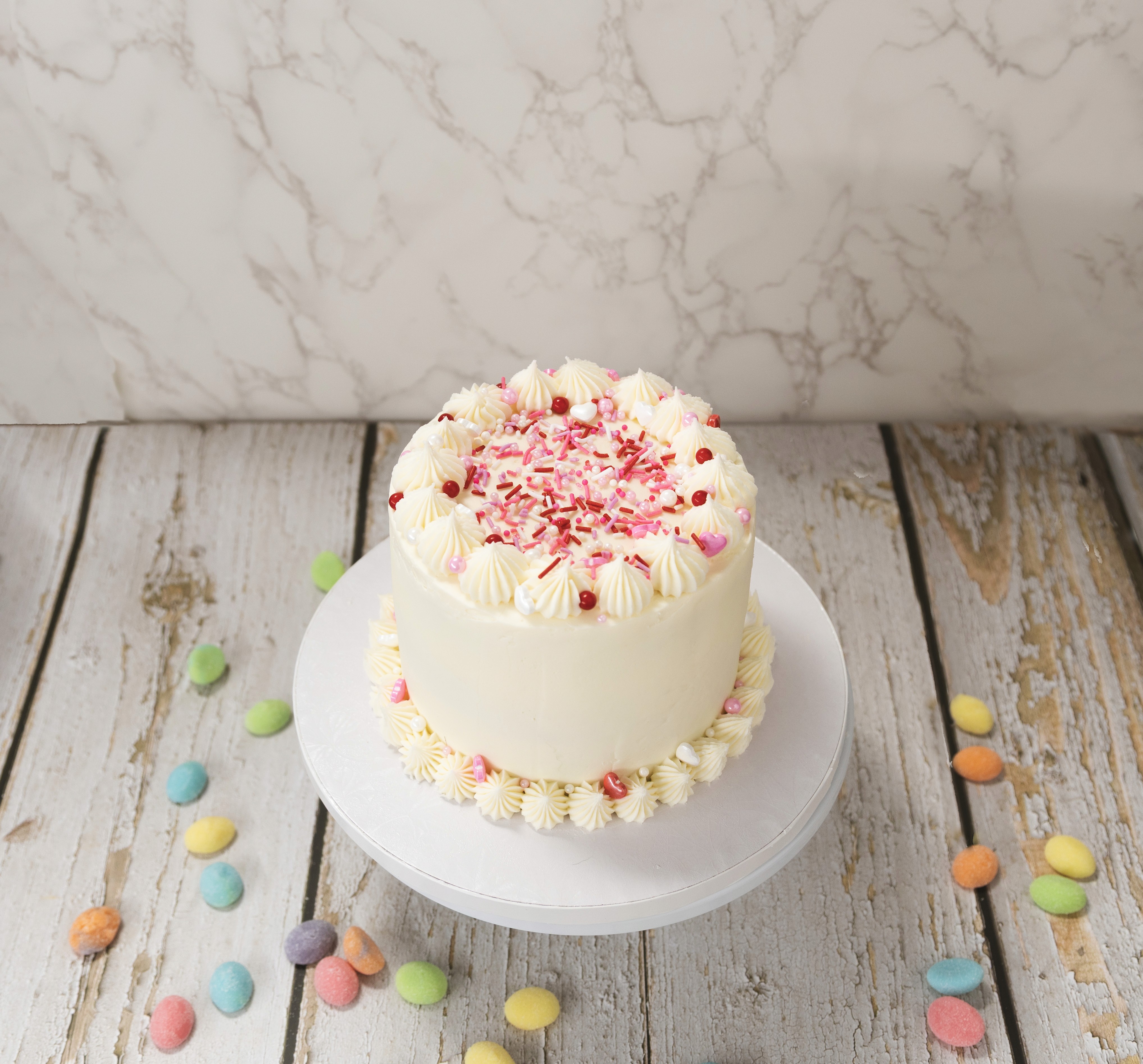 A small white frosted celebration cake topped with pink and red sprinkles, positioned on a minimal white plate over a light wood surface.
