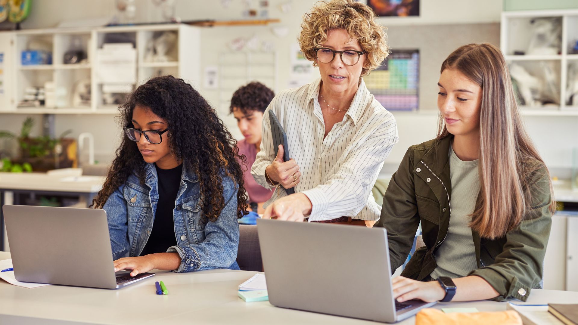 A teacher leans in to guide students working at a laptop in class