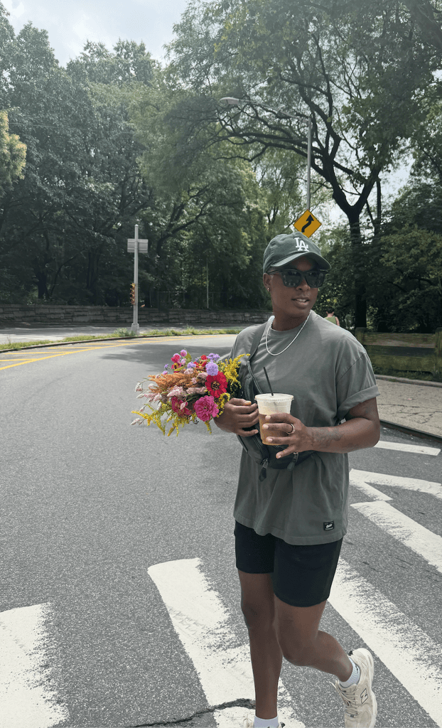 Ashleigh Warren walks along a crosswalk holding colorful flowers and a drink, surrounded by trees and sunny weather.