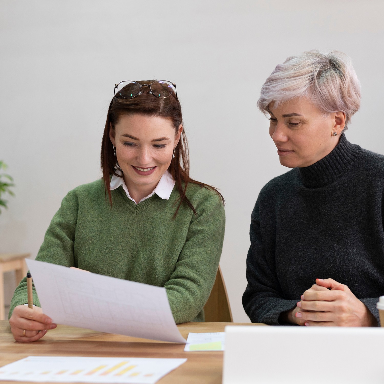 Two women are sitting at a table reviewing documents with graphs and charts, appearing focused and engaged in a collaborative meeting.