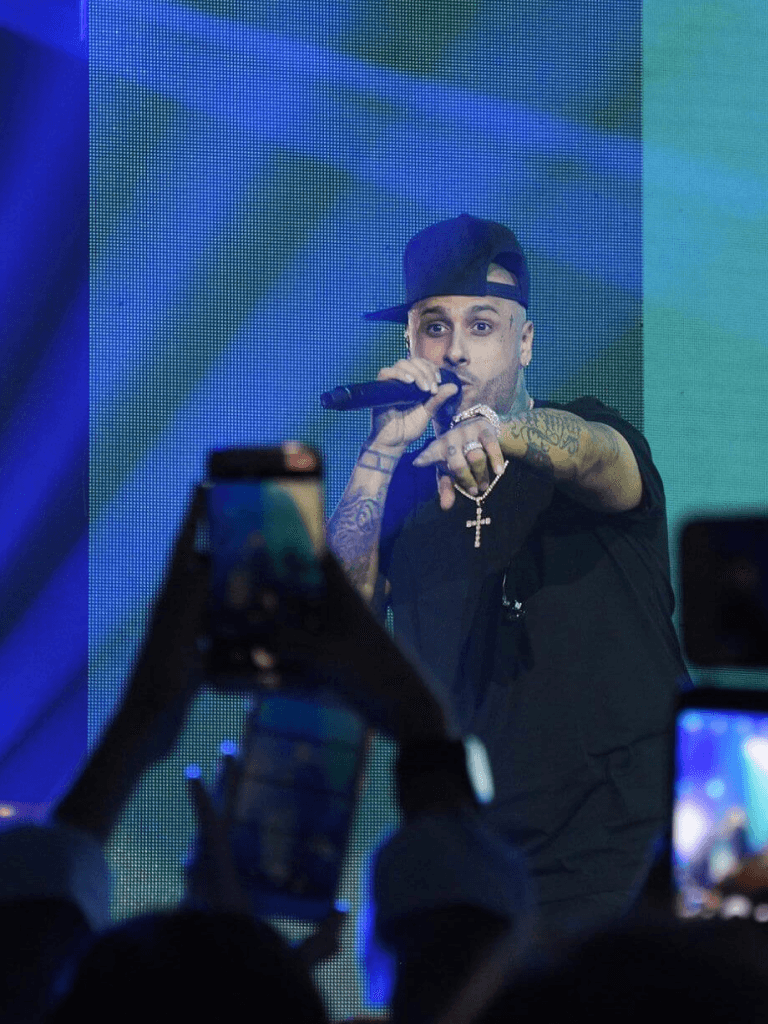 Cantante tatuado con gorra girada canta y señala al público desde escenario azul; asistentes alzan celulares para grabar.