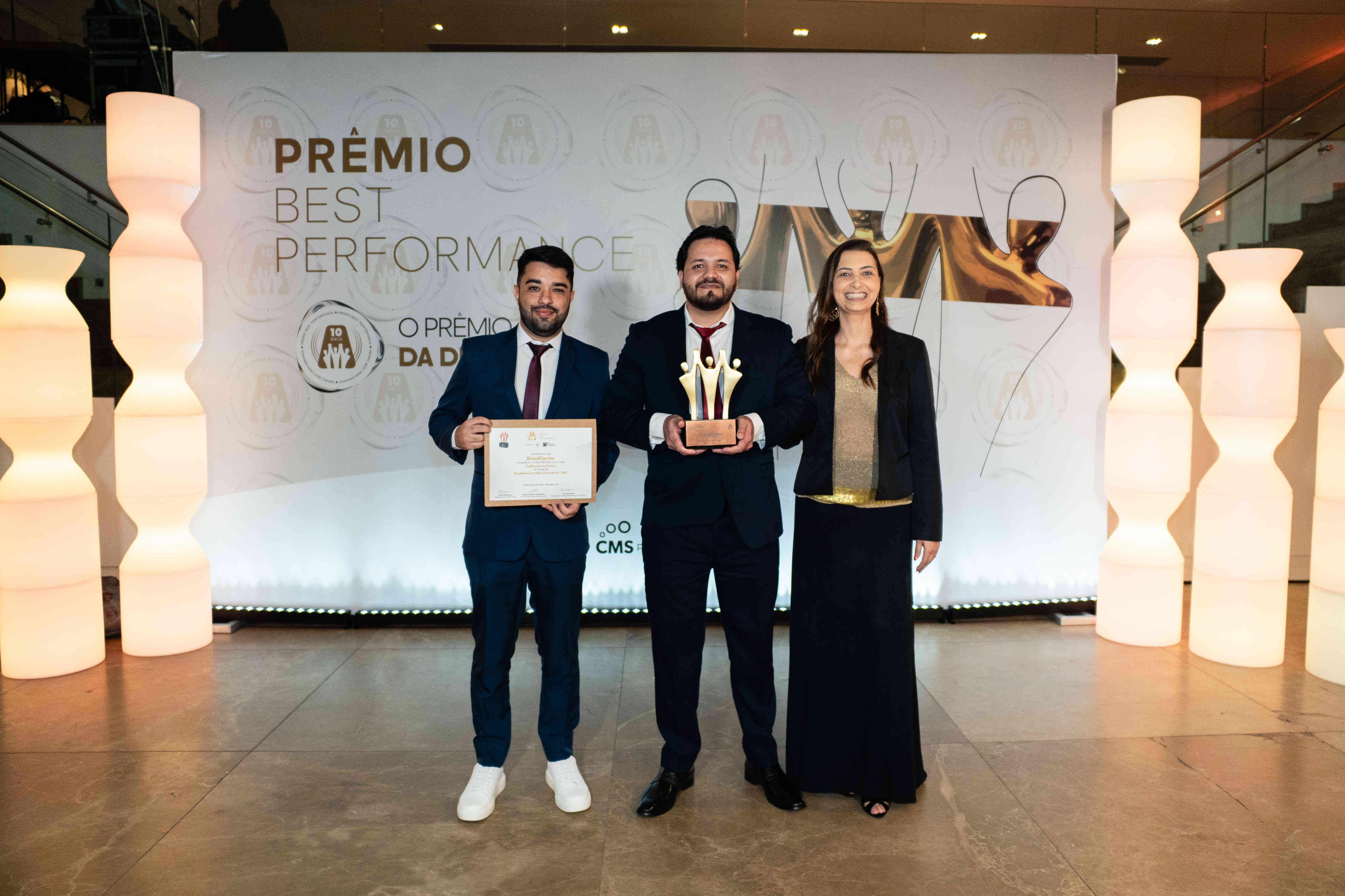 Executivos da BrasilCenter posando com troféu bronze e certificado no palco do Prêmio Best Performance 2025