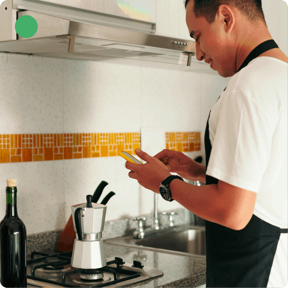 Man in an apron stands at a stovetop using a smartphone while cooking with a moka pot