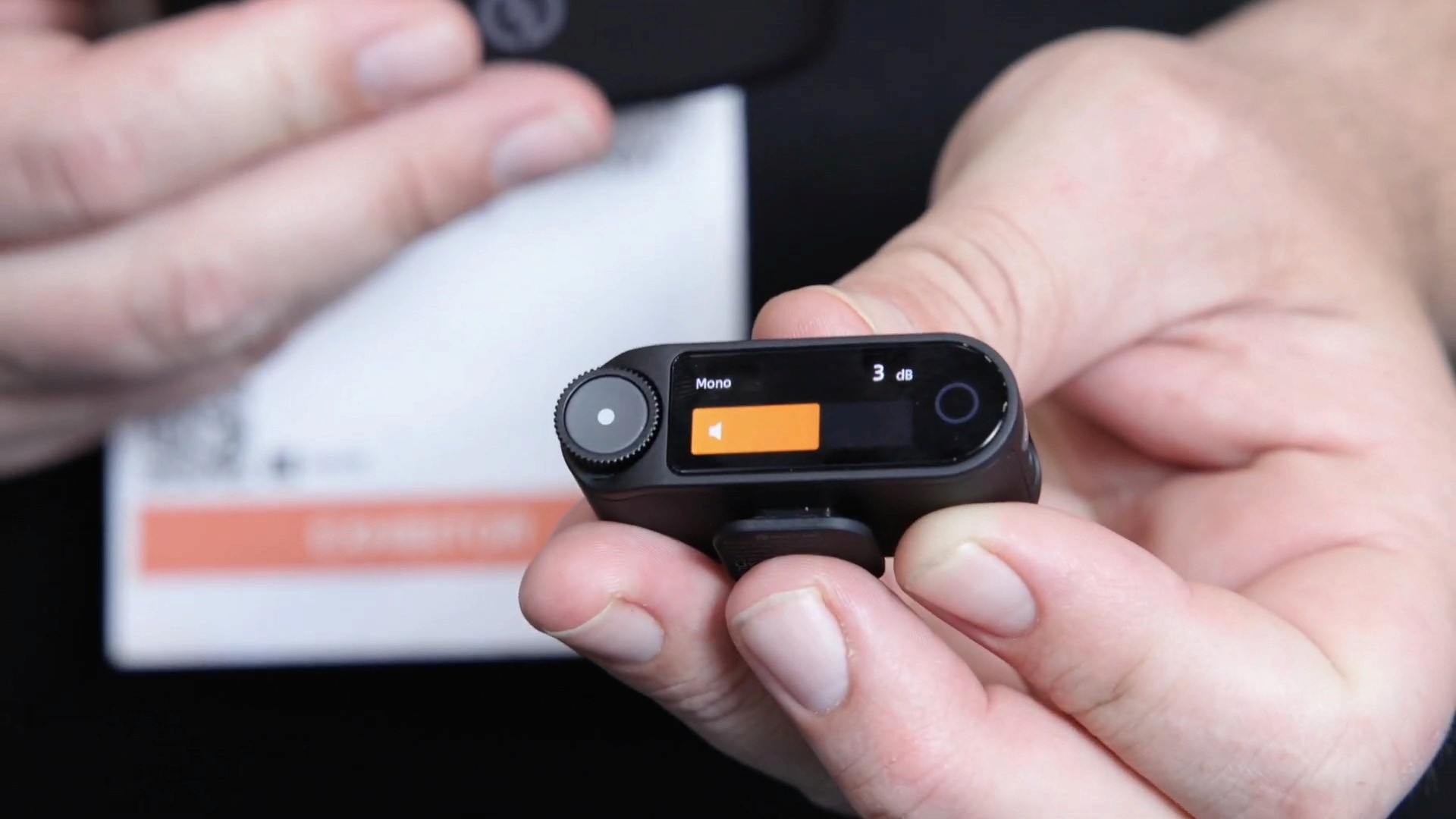 Close-up of hands holding a small, round wireless microphone transmitter with an orange display screen.