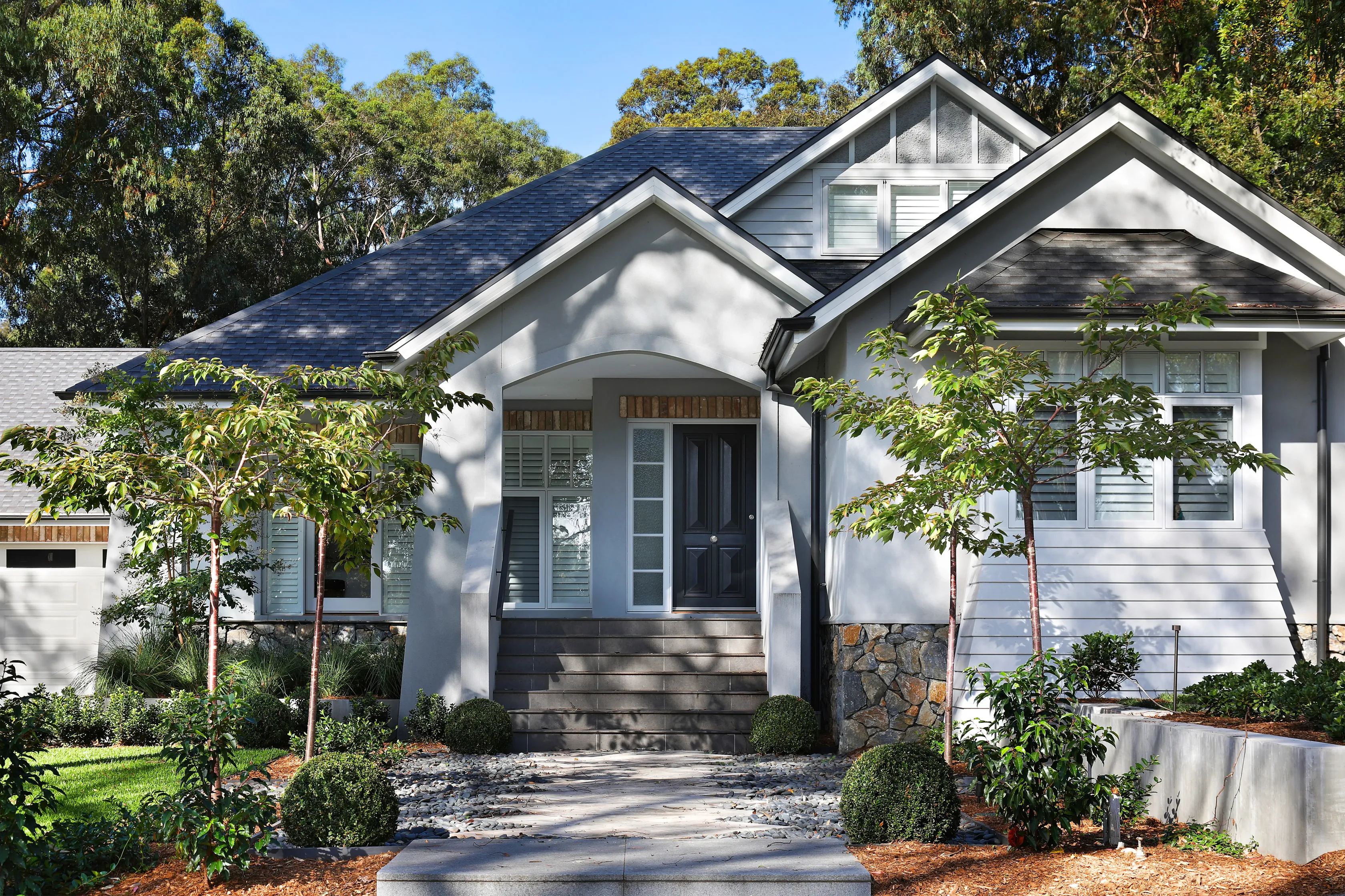 Entry of the Montmorency Residence with a rendered and weatherboard facade, stacked-stone base, steep grey shingle roof, plantation-shutter windows, a dark timber door and a pebble-bordered path through young trees