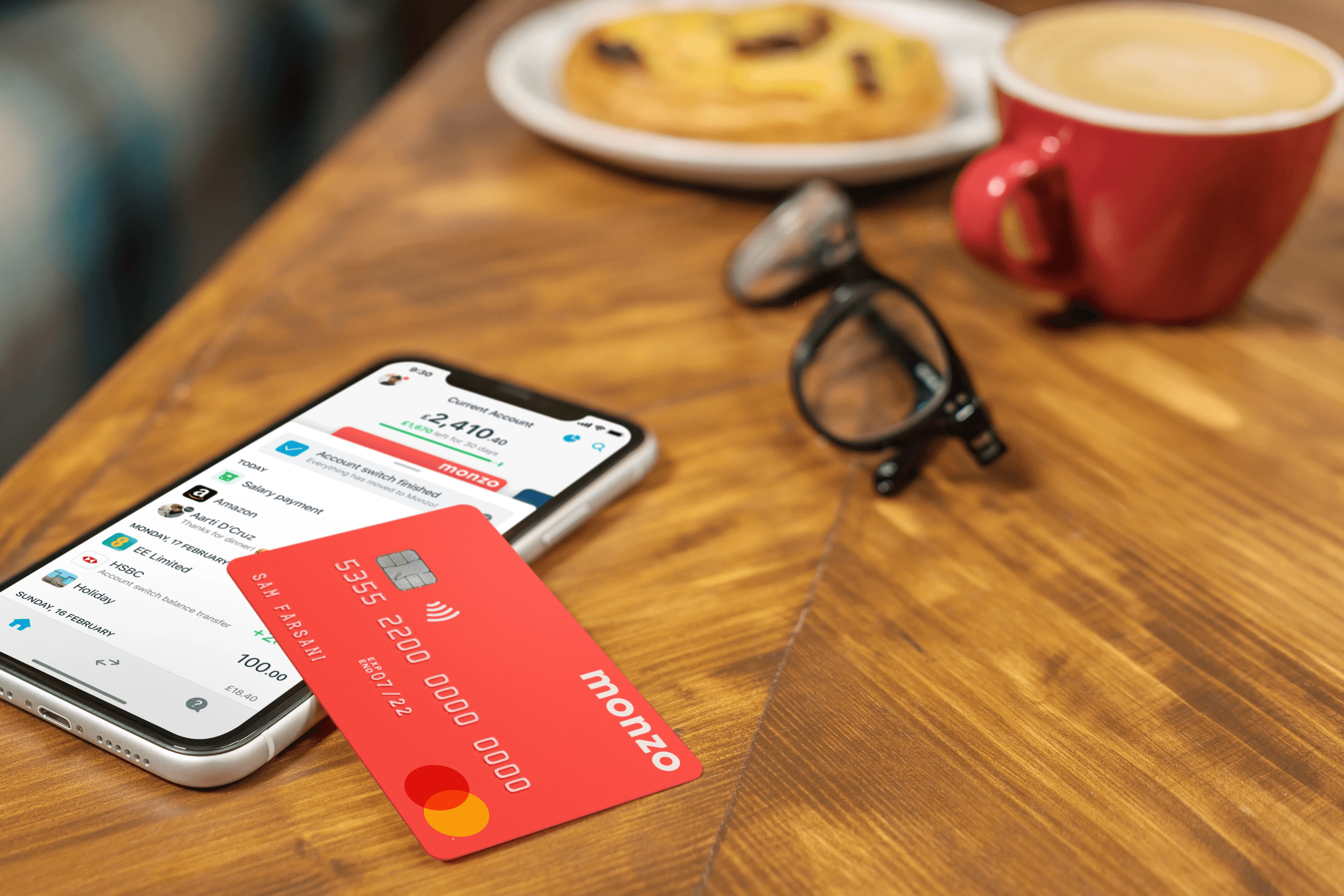 Image of a mobile phone on a table with a Monzo account showing on the screen, and a Monzo bank card on top of the phone at an angle. The setting is in a café, with a coffee, cookie and glasses on the table near the phone and bank card.