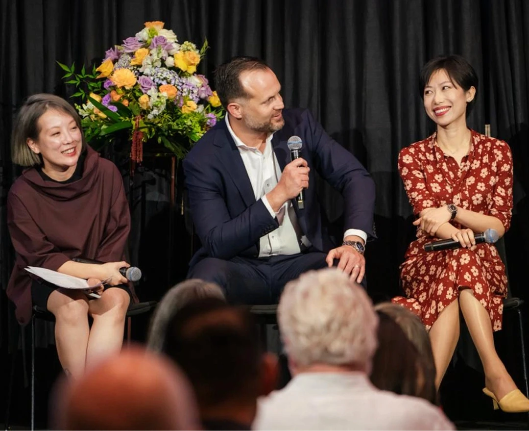 A group of three people sitting on chairs, engaged in a lively discussion with microphones in hand against a backdrop of a vibrant floral arrangement and dark curtains, suggesting a formal event or conference setting.
