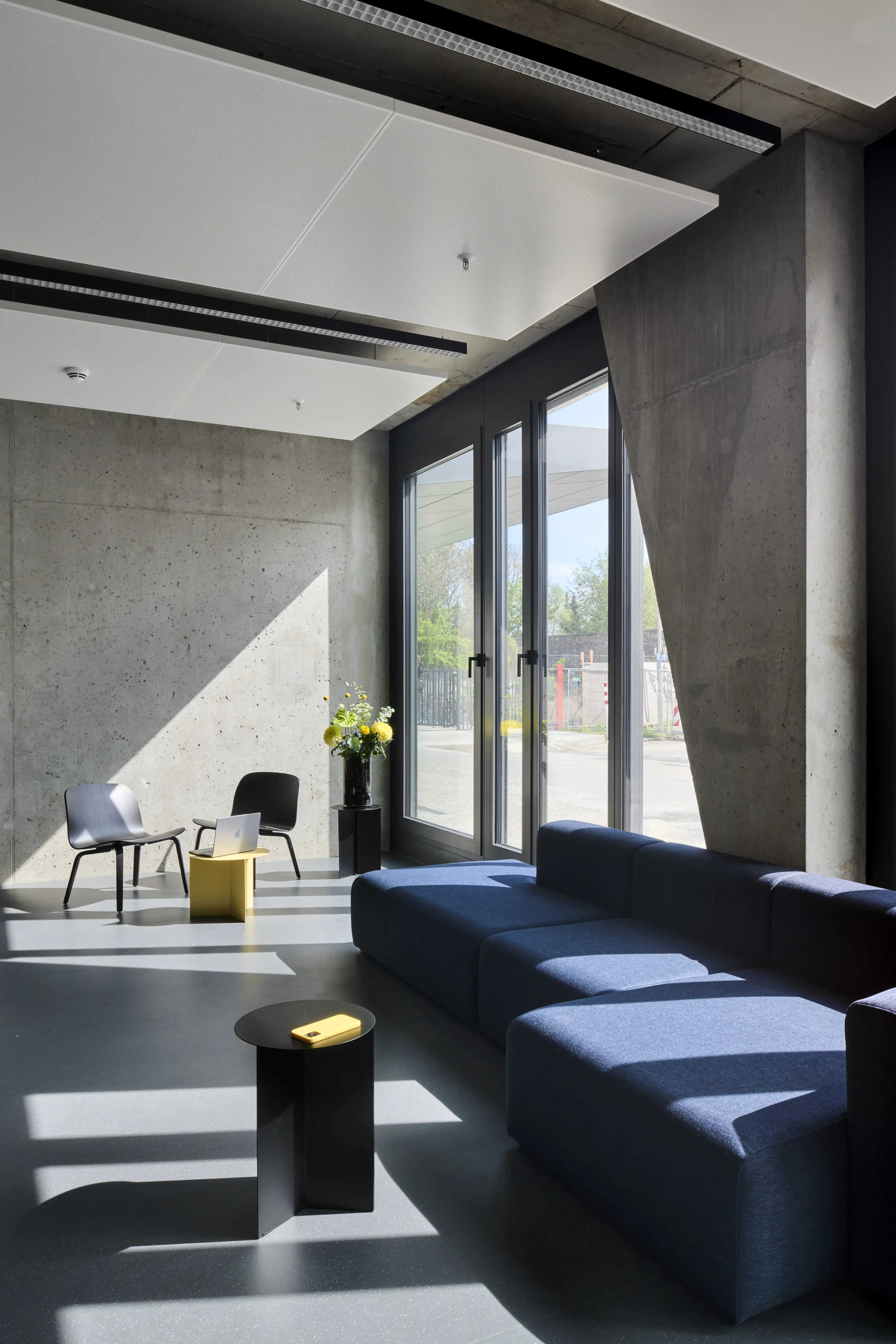Lounge corner at ENPAL with a navy modular sofa, a black round side table, grey lounge chairs, a yellow side table and raw concrete walls by tall windows.