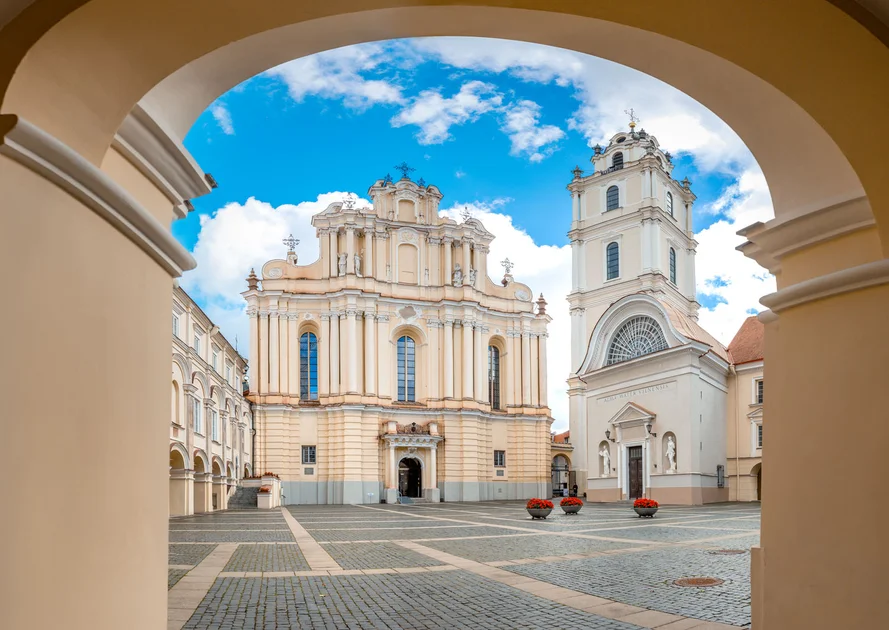 Outdoor restaurant terrace overlooking a tranquil lake at sunset. White tables and chairs are set with glassware. Sheer curtains add elegance. Calm ambiance.