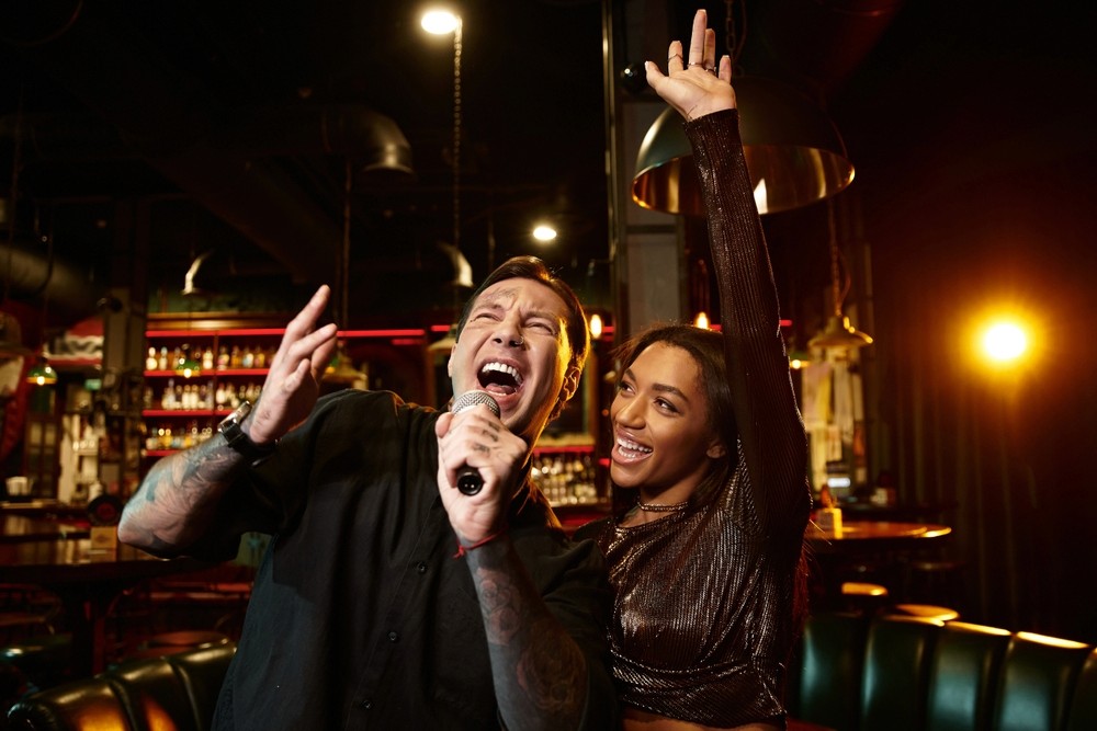 A young person with curly hair smiles at the camera while sitting in front of a laptop against a dark background.
