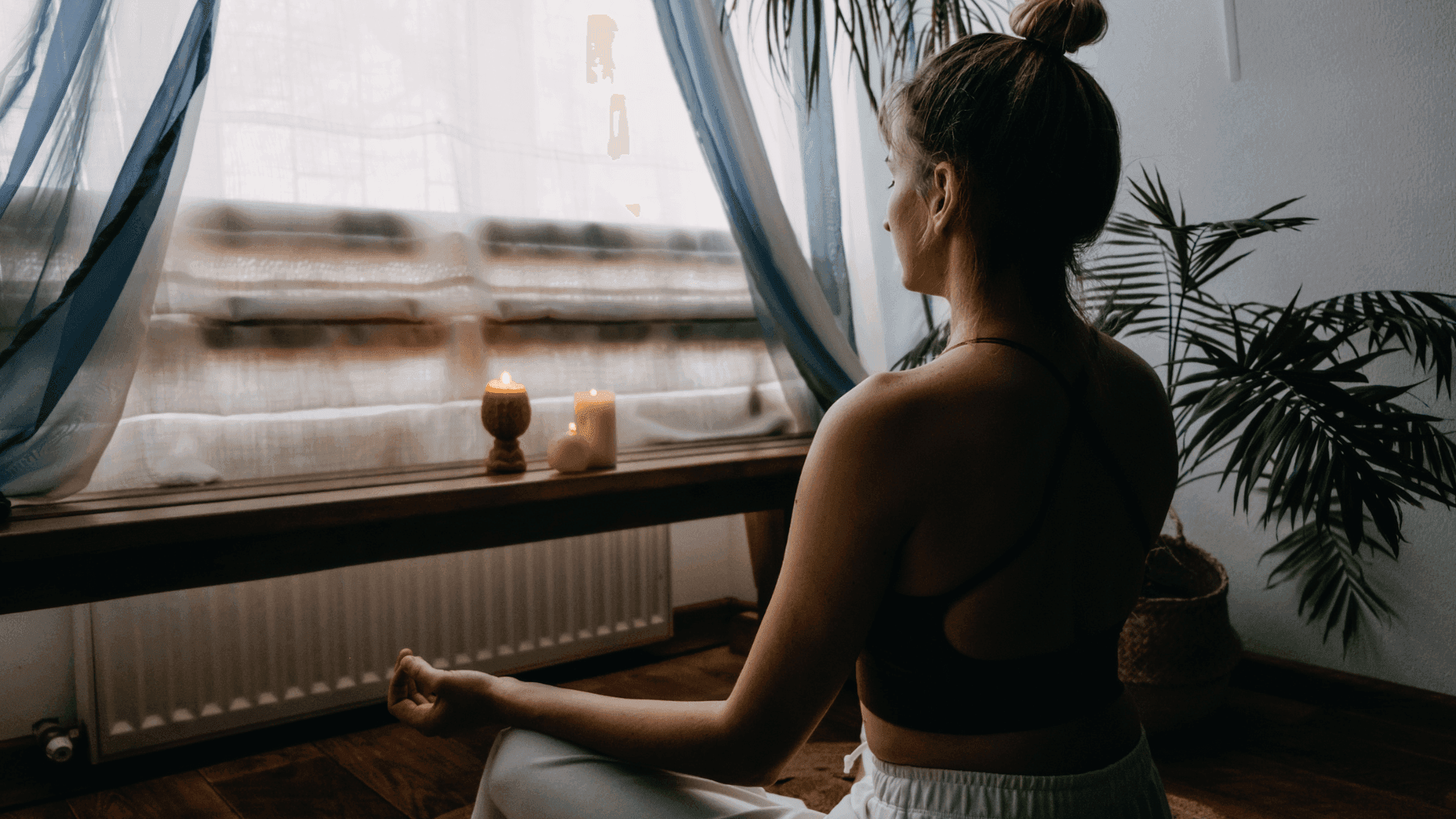 Woman practicing meditation in Easy Pose (Sukhasana) by window with lit candles, wearing yoga attire, creating a peaceful home meditation space with palm plants and soft curtains