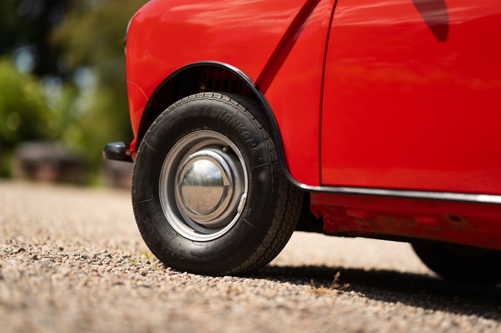 A close-up of a red car's wheel resting on a gravel road with greenery in the background.