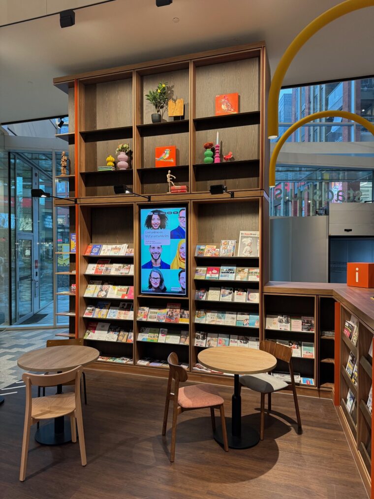 Modern library interior featuring a vertical digital signage display embedded in wooden bookshelves, used for promoting library content, events, and community information alongside magazines and reading areas.