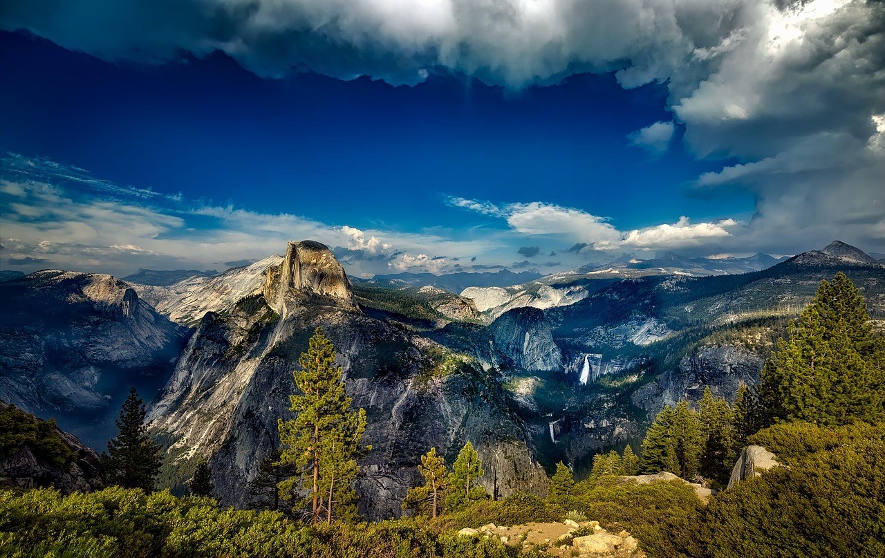 Dramatic Yosemite National Park landscape with Half Dome peak, Yosemite Falls, and dense pine forests under a cloudy blue sky.