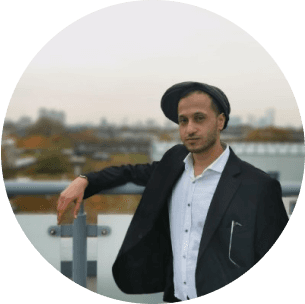 A person in a black suit and white shirt stands on a rooftop with a cityscape in the background, wearing a casual cap and leaning on a metal railing.