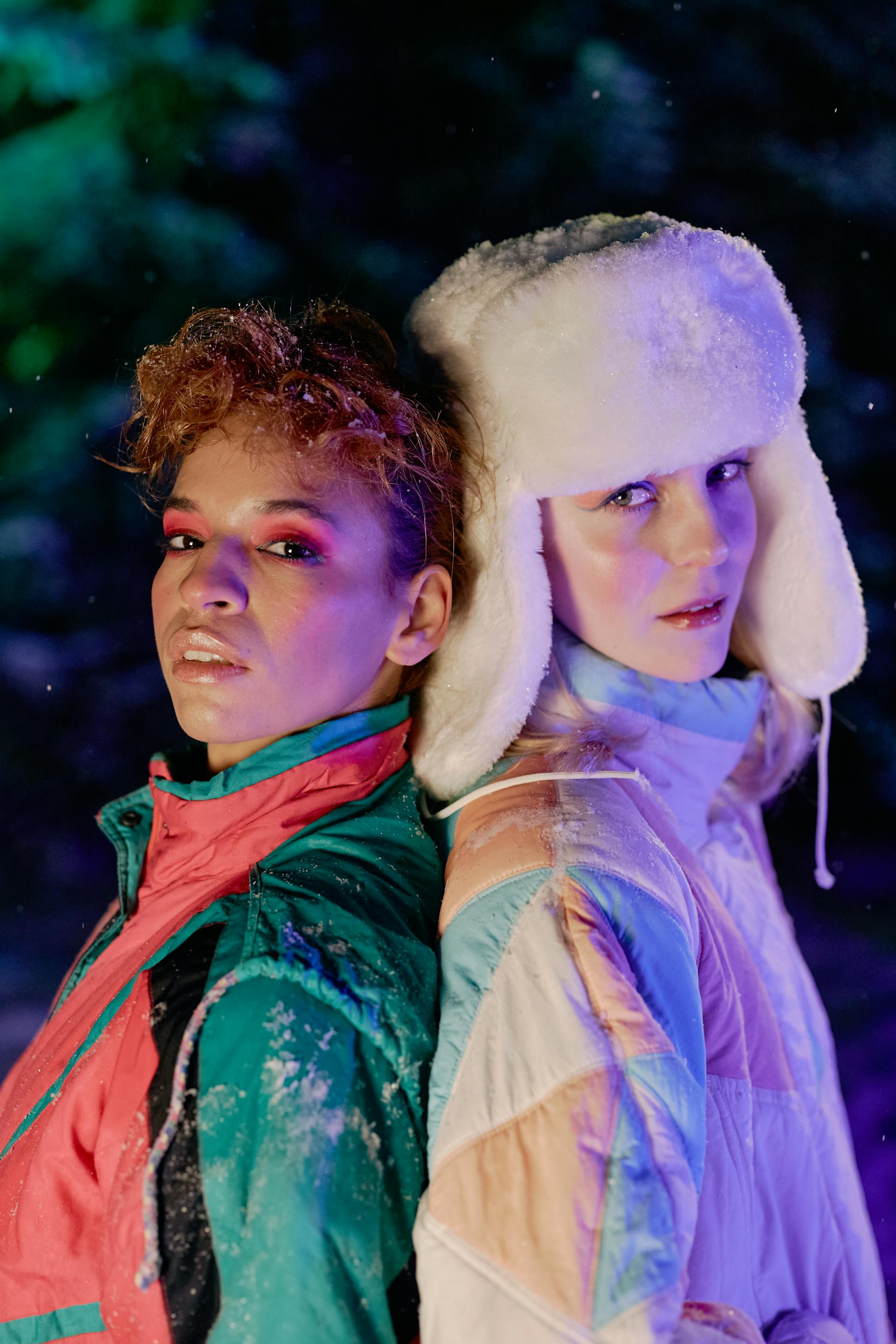 A close-up portrait of two people standing back-to-back in the snow; one has curly hair and bold pink eyeshadow, and the other wears a white faux-fur ushanka hat.