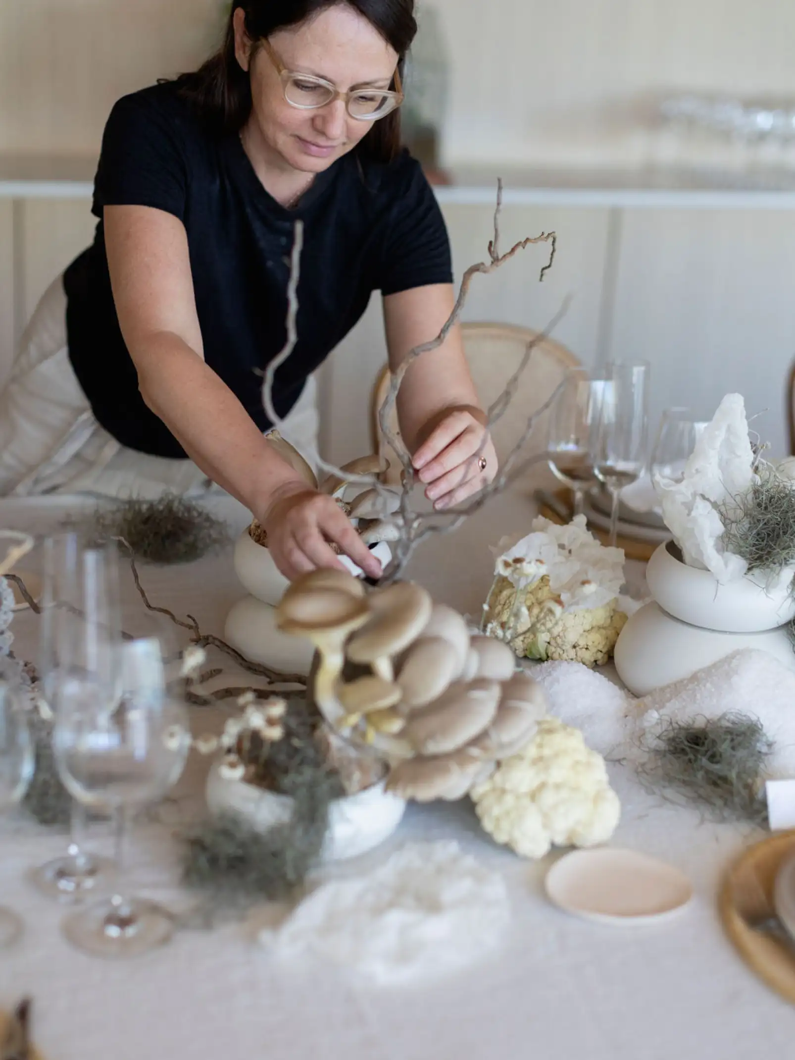 Irina Khatsernova armando una decoración de mesa con hongos ostra, ramas secas y elementos naturales sobre un mantel blanco.