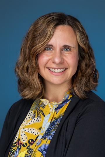 Smiling woman with shoulder-length brown hair, wearing a patterned top and black cardigan, in a professional portrait.
