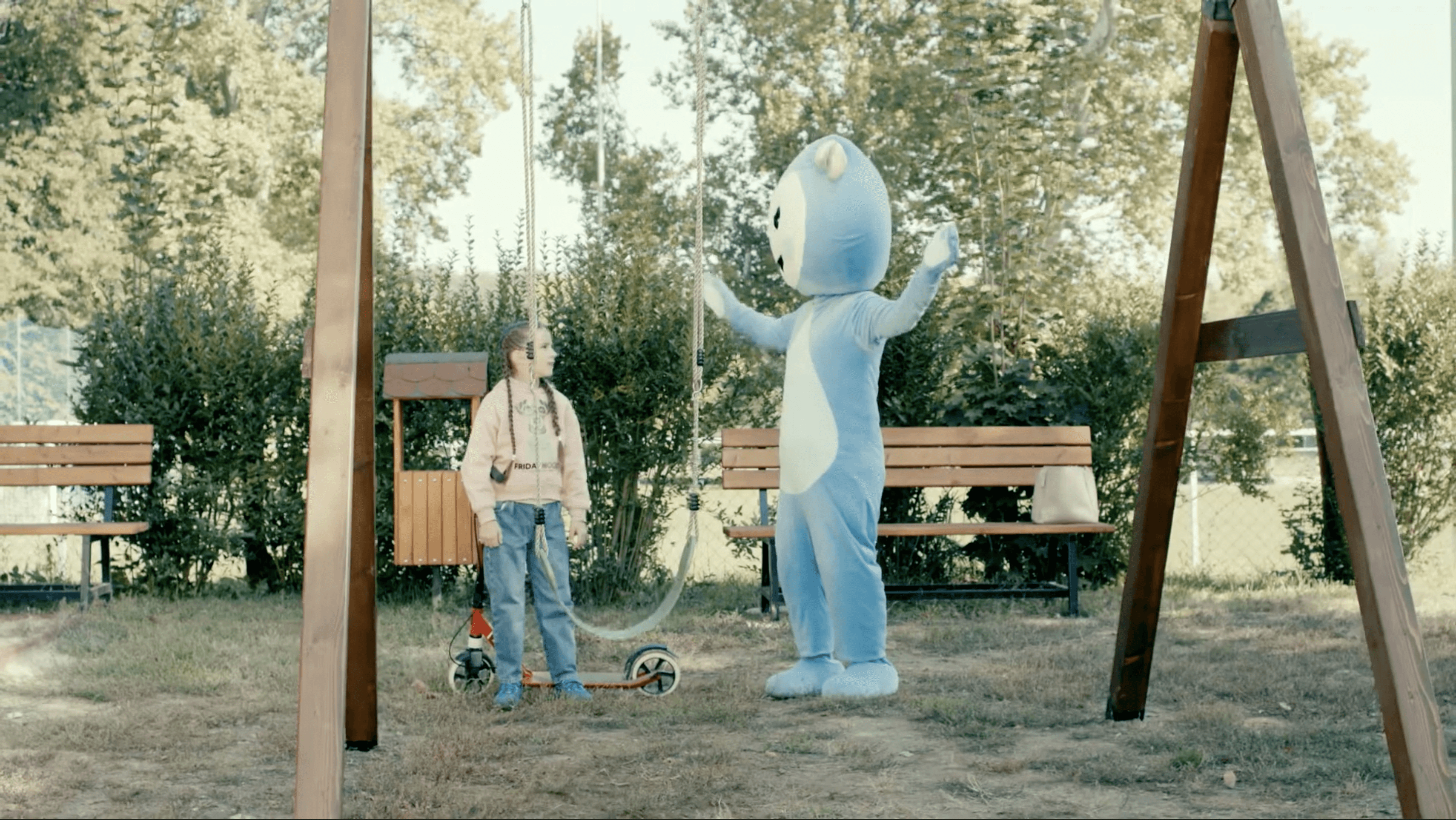 Cseppbarát kék kabalafigurája a játszótéren egy hinta melett a rollerező kislánnyal.