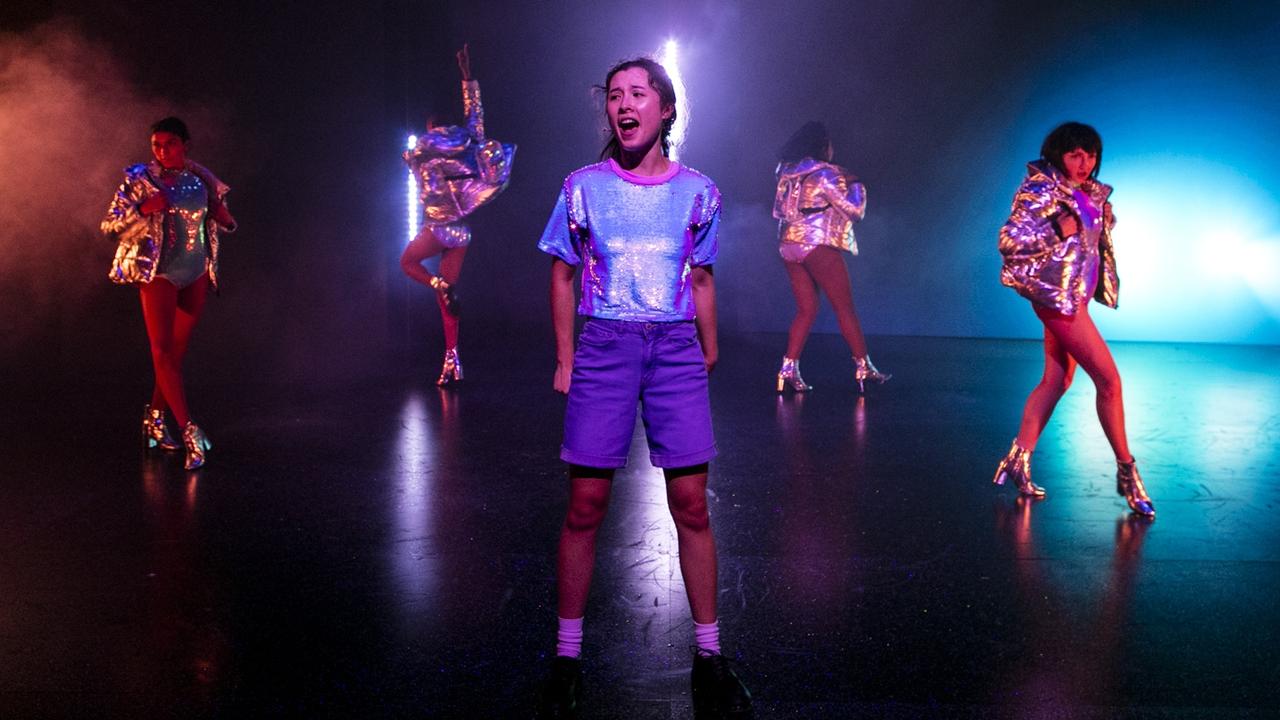 A central performer sings in a shimmering purple costume while four dancers in metallic jackets and boots strike bold poses in a smoke-filled, neon-lit scene.