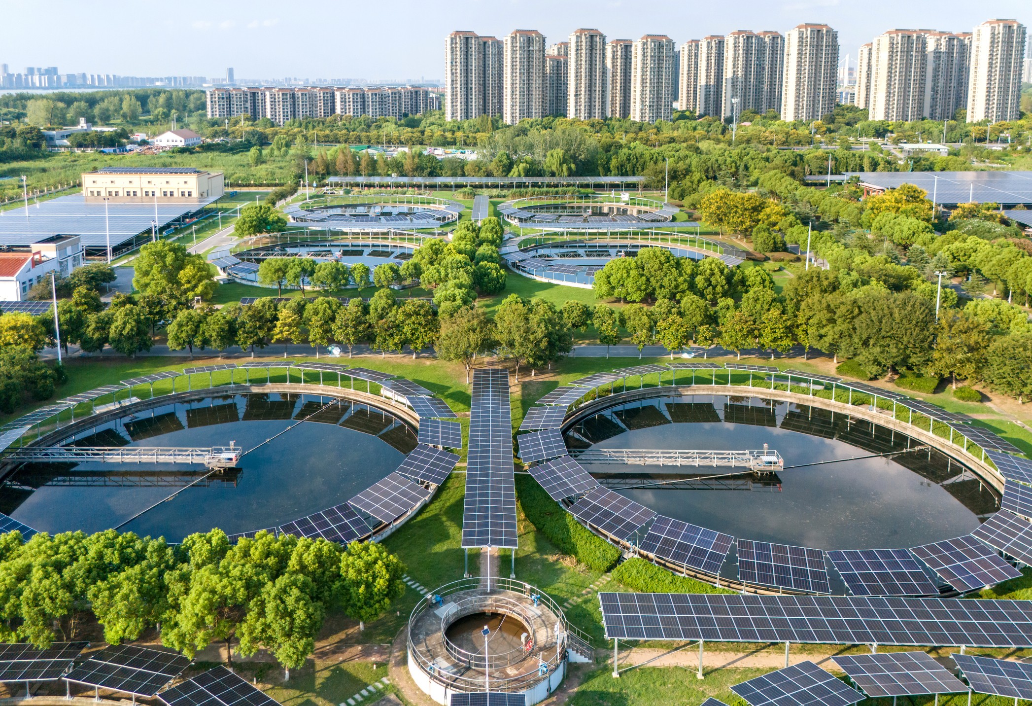 Aerial view of a landscaped area with ponds and modern buildings in the background, surrounded by green trees.