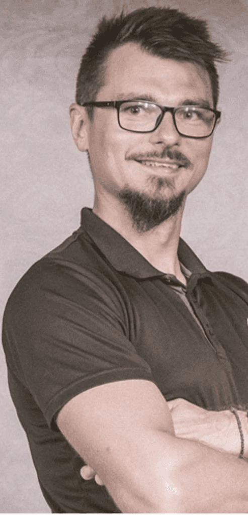 Zdjęcie przedstawia kobietę o jasnych, falowanych włosach i okularach w ciemnych oprawkach. Ma na sobie czarną bluzkę z wzorem i patrzy prosto w obiektyw. Tło jest jednolite, ciemne.