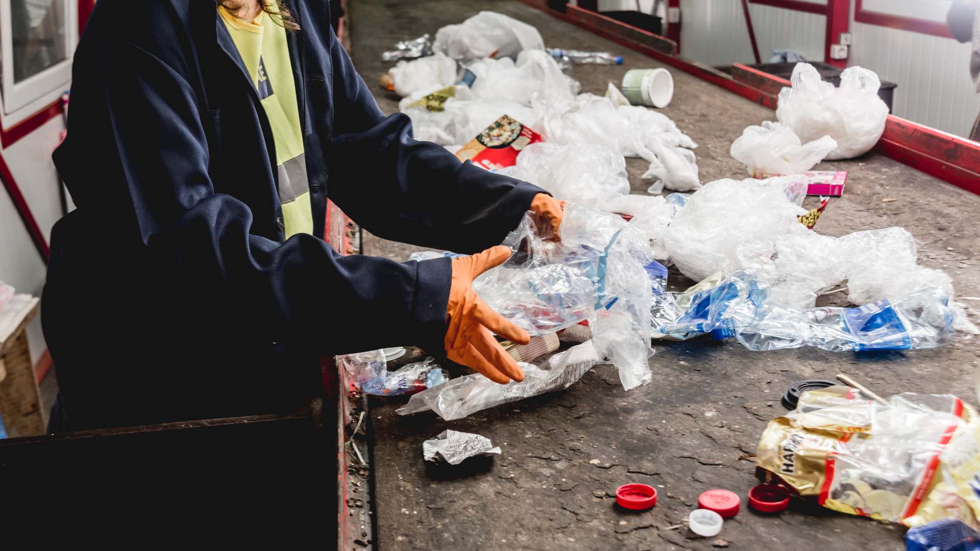 Worker in a dark coat and orange gloves sorting plastic packaging waste on a conveyor at a recycling facility