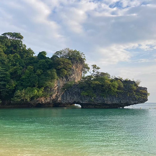 岩石岛上覆盖着茂密的绿色植物,自然拱门悬于碧绿色的海水之上,天空部分云层笼罩。