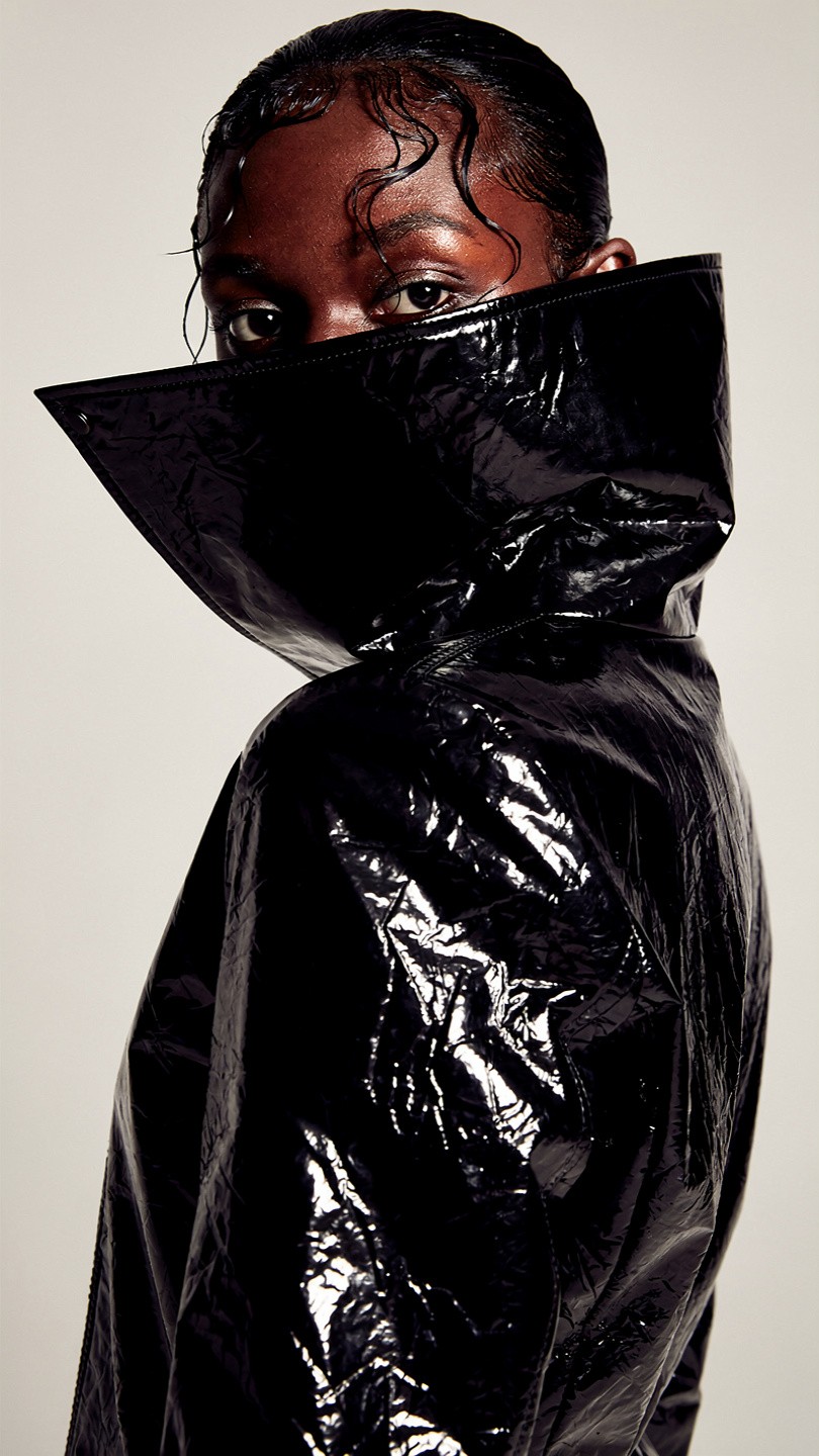A person wears a shiny black vinyl coat with a high collar, creating a dramatic and modern look. The expression is confident and bold against a neutral background.