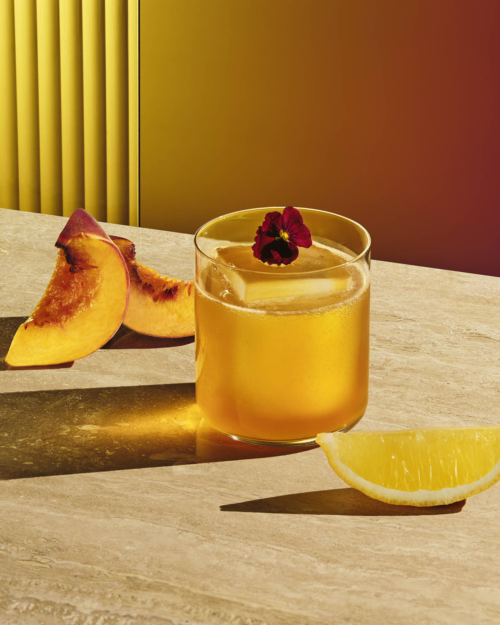 A glass of yellow cocktail topped with a flower, beside fresh peach slices, on a wooden table with a gradient background.