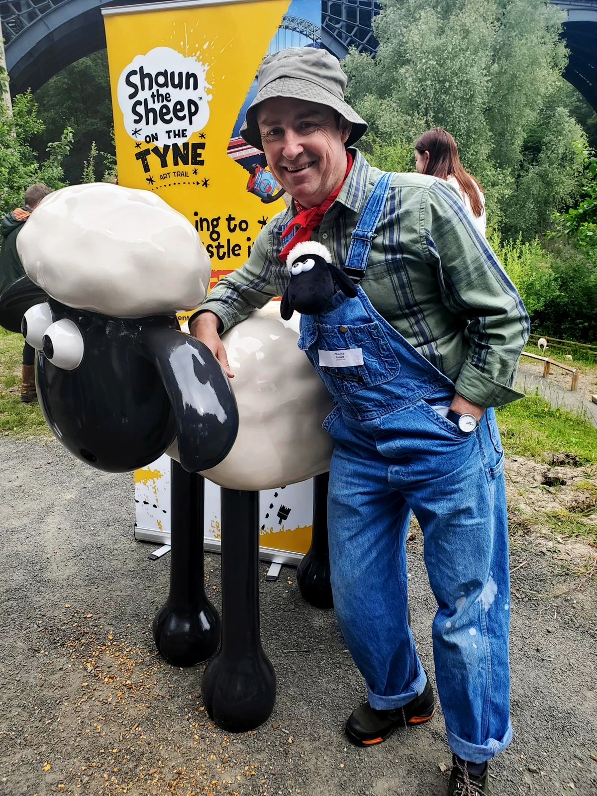 A person dressed in overalls, a checkered shirt, and a floppy hat poses beside a large Shaun the Sheep sculpture, with a bright yellow “Shaun the Sheep on the Tyne” art‑trail sign in the background.