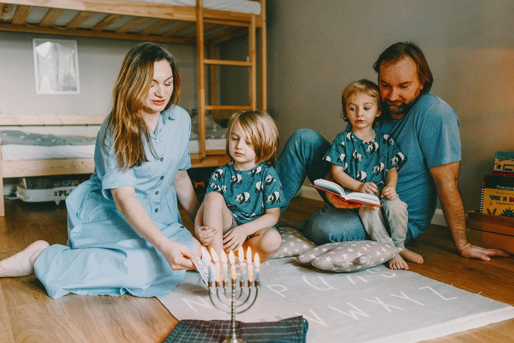 A family of four celebrates Hanukkah at home, gathered around a lit menorah. The mother, dressed in a light blue dress, lights the candles, while the father, wearing a blue shirt, reads a book to their two young children, who are dressed in matching blue pajamas with dog prints. The scene is warm and intimate, highlighting the family's togetherness during the holiday.