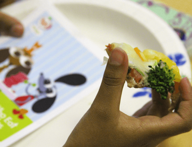 A child's dark-skinned hand holds a piece of food that appears to be an omelet or frittata. The food has visible pieces of broccoli and tomato and is topped with melted cheese. The hand is positioned over a white plate with a decorative design featuring blue and green floral patterns. To the left of the plate is a sheet with cartoon characters, including a character resembling Mickey Mouse with large round ears and a red bow-tie-like element.