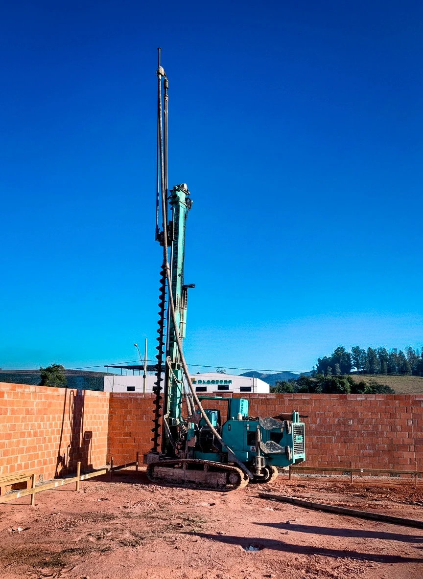 Máquina perfuratriz alta em primeiro plano, com o mastro totalmente estendido, em um terreno de terra vermelha. Ao lado, um muro de tijolos aparentes, sob um céu azul vibrante.