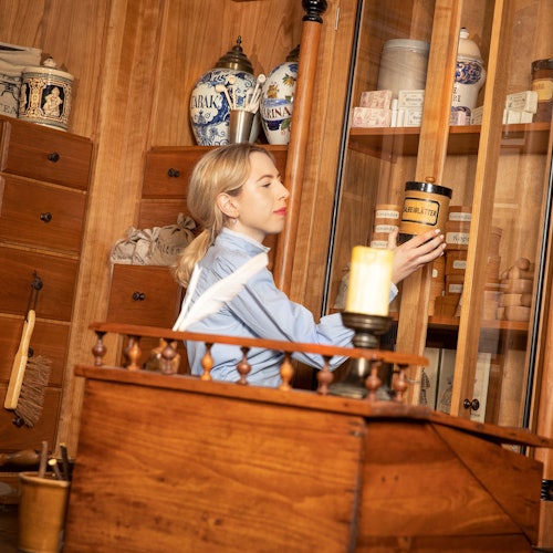 A woman in a blue shirt arranges jars in a wooden cabinet filled with apothecary items in a vintage-looking room.