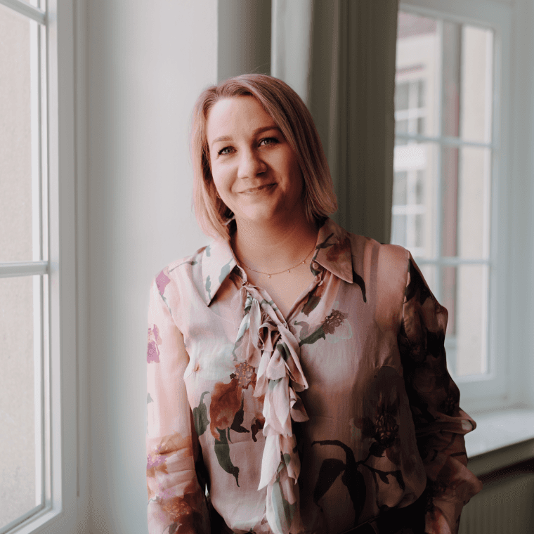 Jüngere Geschäftsführerin mit blumiger Bluse steht vor einem Fenster im Büro von SLB und blickt in die Kamera.