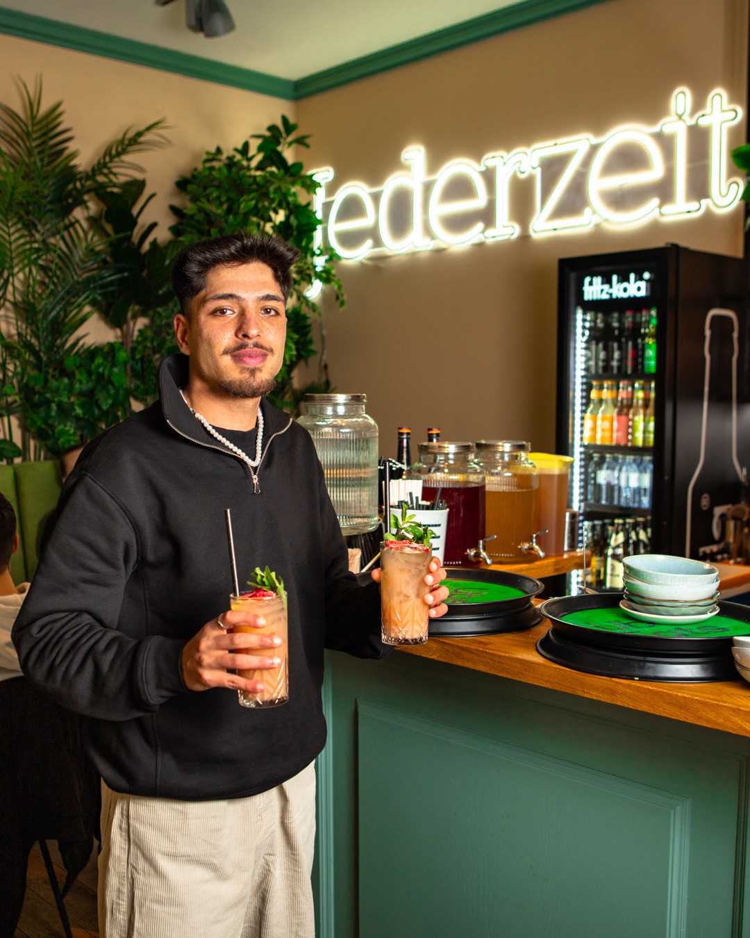 Ein Mann hält zwei farbenfrohe Getränke mit Garnierung in den Händen, er steht vor einer Theke mit einem Neon-Jederzeit-Schild und einem Kühlschrank.