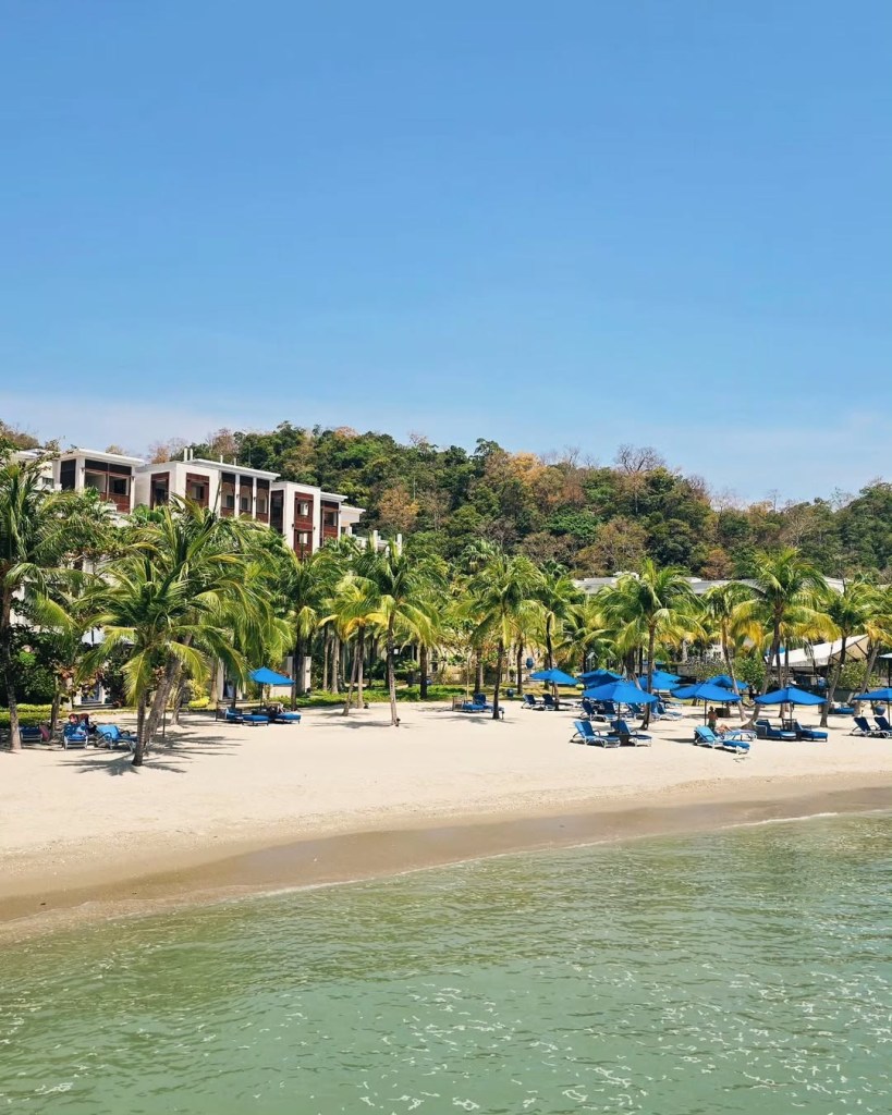 View of The St. Regis, Langkawi beach with palm trees and blue lounge chairs by the water.
