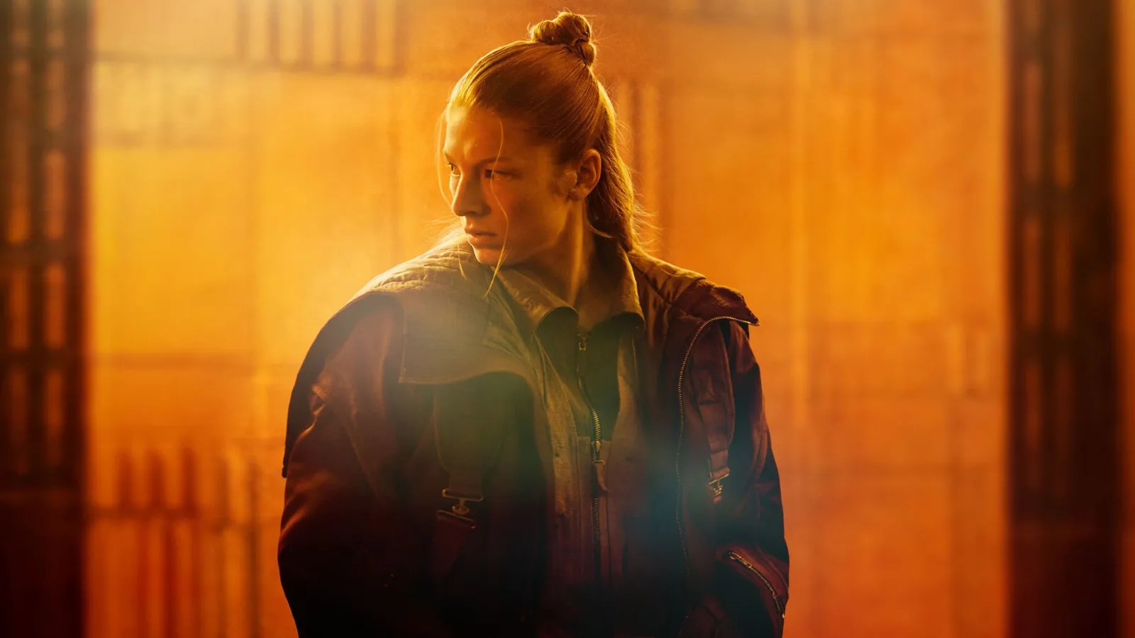 A young woman with a top knot hairstyle is illuminated by warm, golden light, wearing a dark jacket with an intense expression, set against a blurred, architectural background.